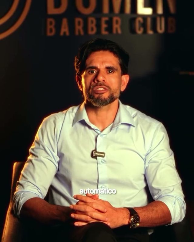 Barbeiro sentado diante do letreiro Domen Barber Club